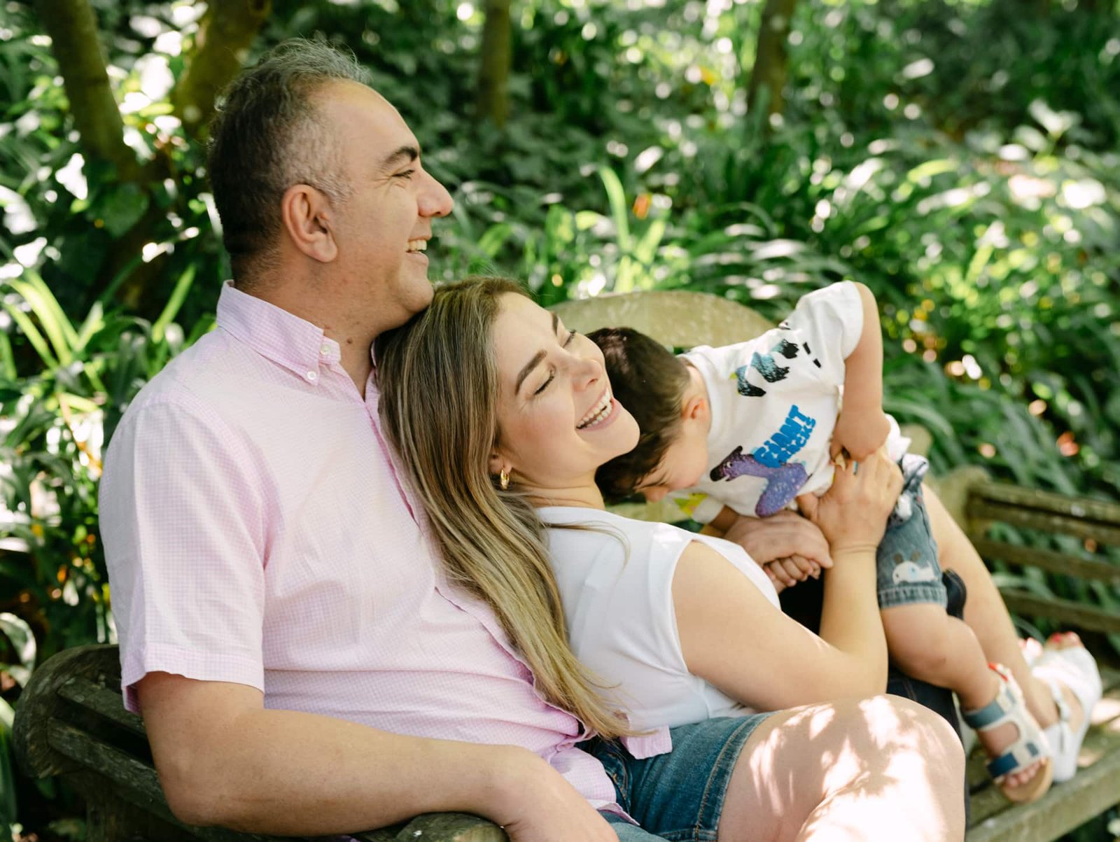 Nasim laying back into Alis front while on a wooden bench and laughing as Adrian plays during a family session at Woodlands Estate