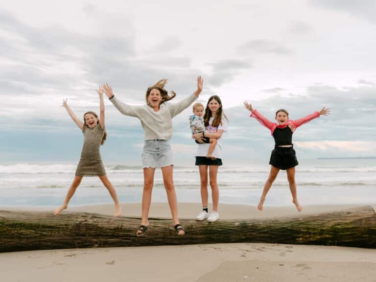 Three girls star jumping off log as the older one holds the young boy during a family session at Papamoa