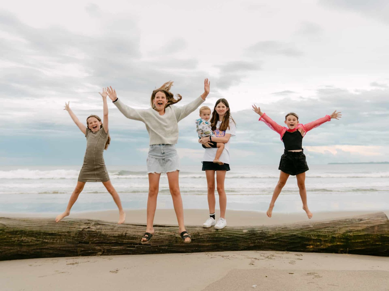 Three girls star jumping off log as the older one holds the young boy during a family session at Papamoa