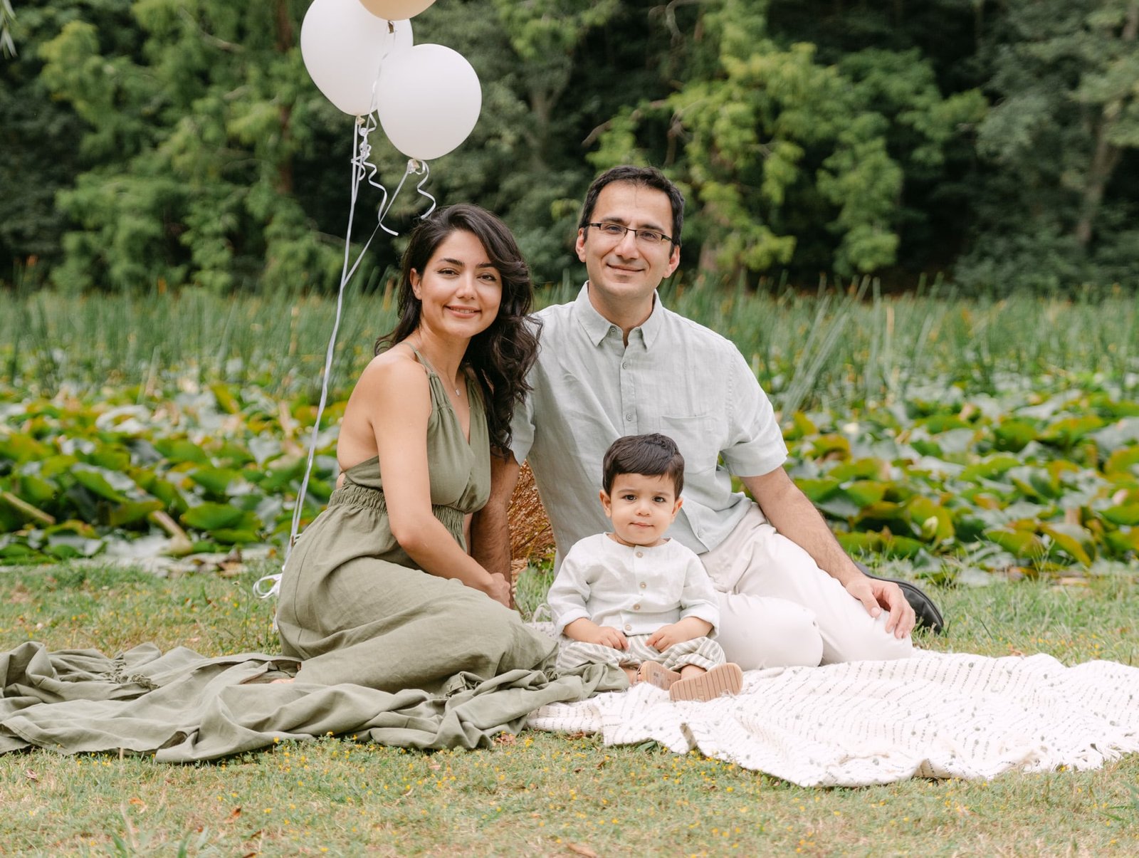 mum dad and little boy sitting on a blanket in a park looking at camera during a family session at Cambridge Lake