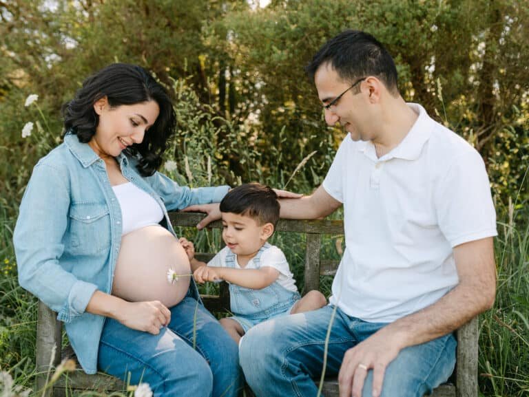 Neil poking Tina's pregnant tummy and Pouya sitting beside him during a maternity session at Hamilton in a park.