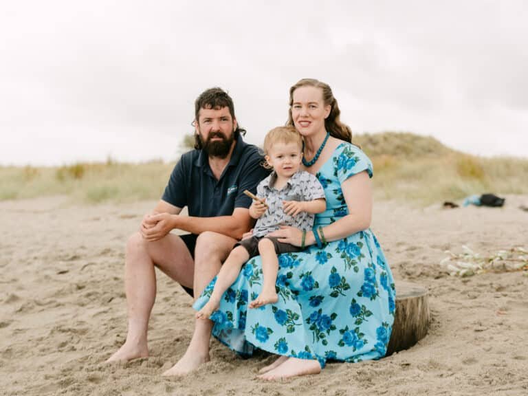 Helen with Will on her lap and Alex sitting next to them, all looking at camera for a photo during a family session at the beach